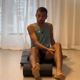 Corredor sentado no chão de casa, sobre um tapete e com rolo de liberação, segurando o pé machucado