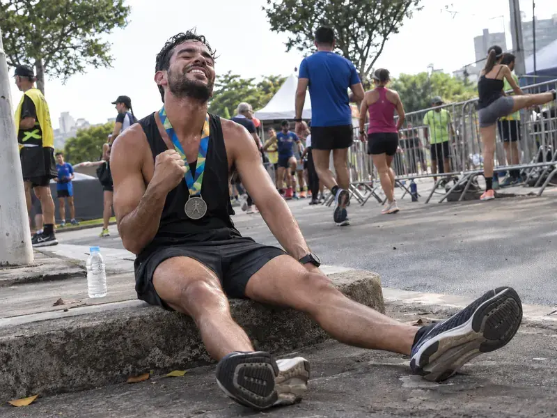 Corredor exausto sentado no meio-fio depois da prova, com a medalha no pescoço, comemorando o próprio tempo