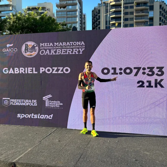Gabriel Pozzo sorrindo com a medalha e apontando para o próprio nome e o tempo de 1:07:33 no painel da Meia Maratona Internacional de Florianópolis